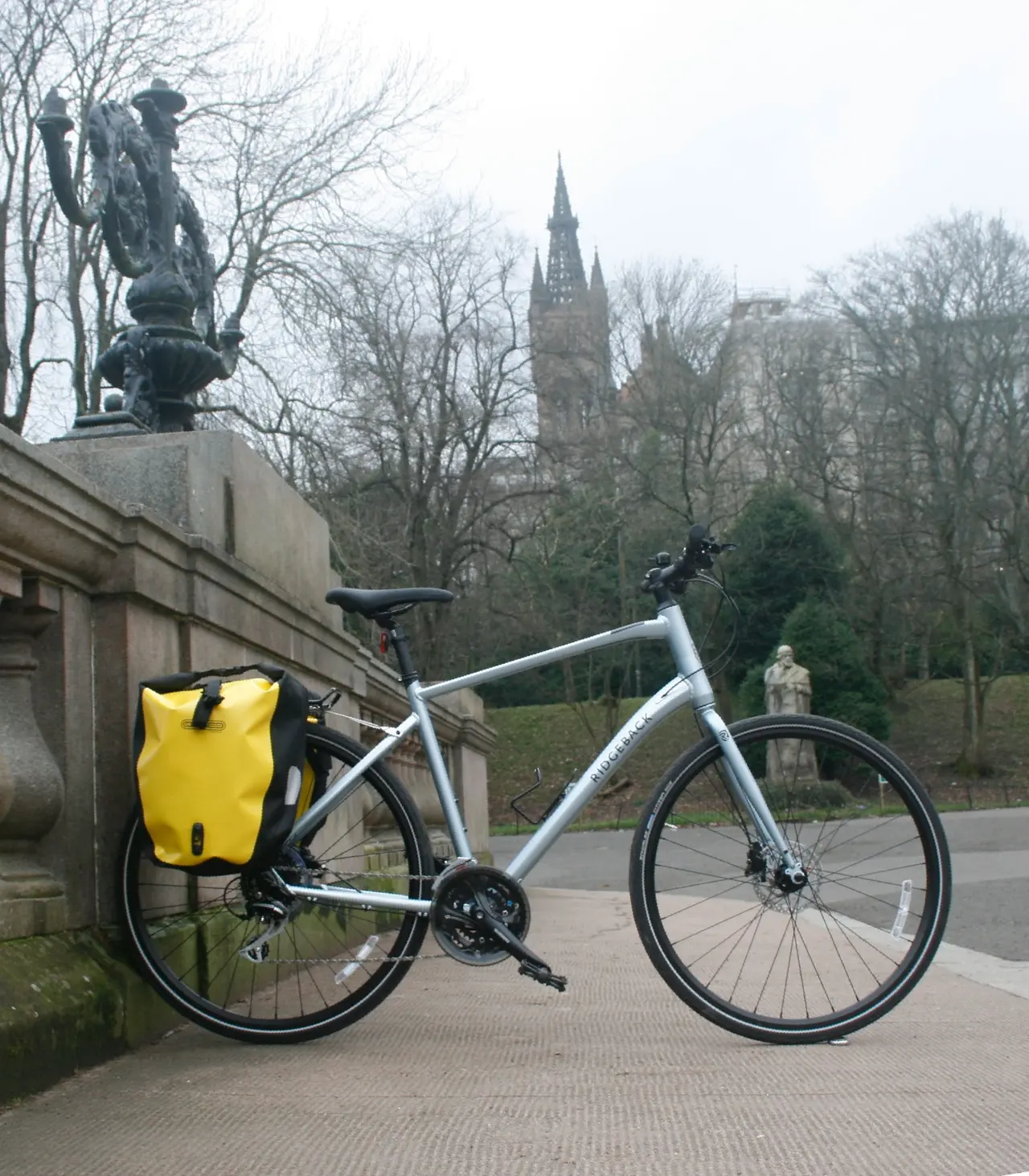 A hire bike with a yellow pannier beside the railings at Kelvingrove Park, with the University tower behind